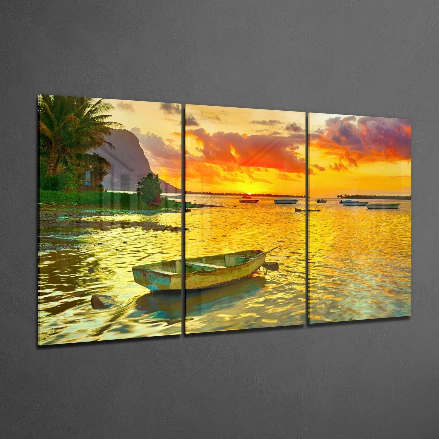 Three-panel glass wall art displaying a tropical sunset beach scene with moored fishing boats, golden water, and palm trees against gray wall.