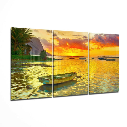 Three-panel glass wall art displaying weathered boats in golden sunset waters with palm trees and dramatic orange sky.