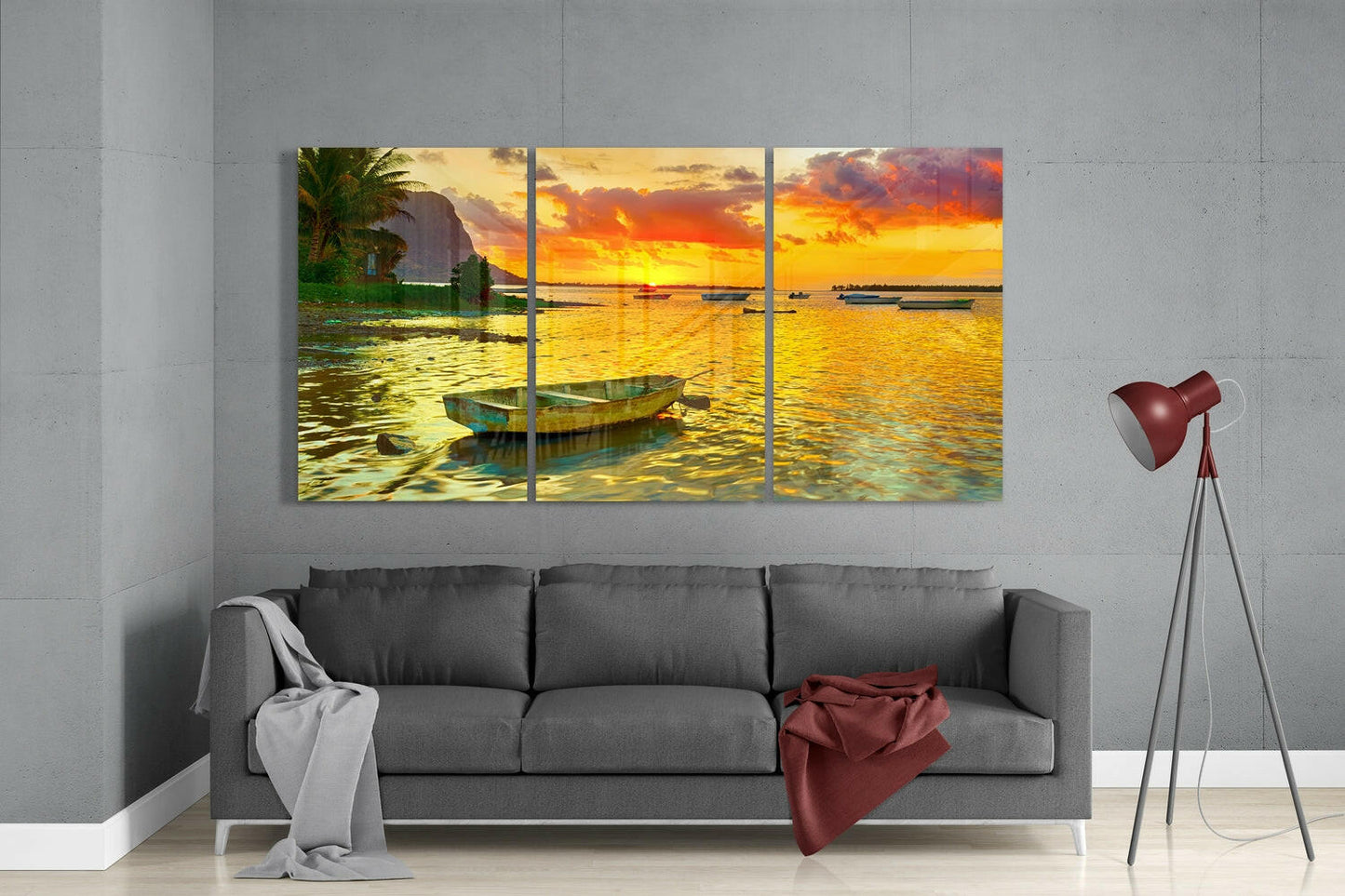 Three-panel glass wall art displaying a golden sunset over calm waters with a moored boat, hung above a gray sofa in a modern living room.