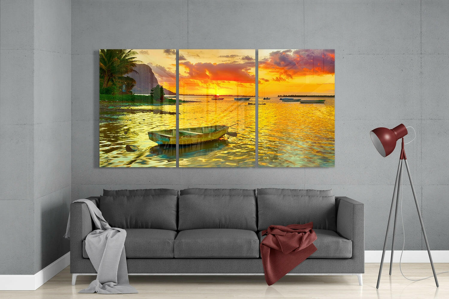 Three-panel glass wall art displaying a golden sunset over calm waters with a moored boat, hung above a gray sofa in a modern living room.