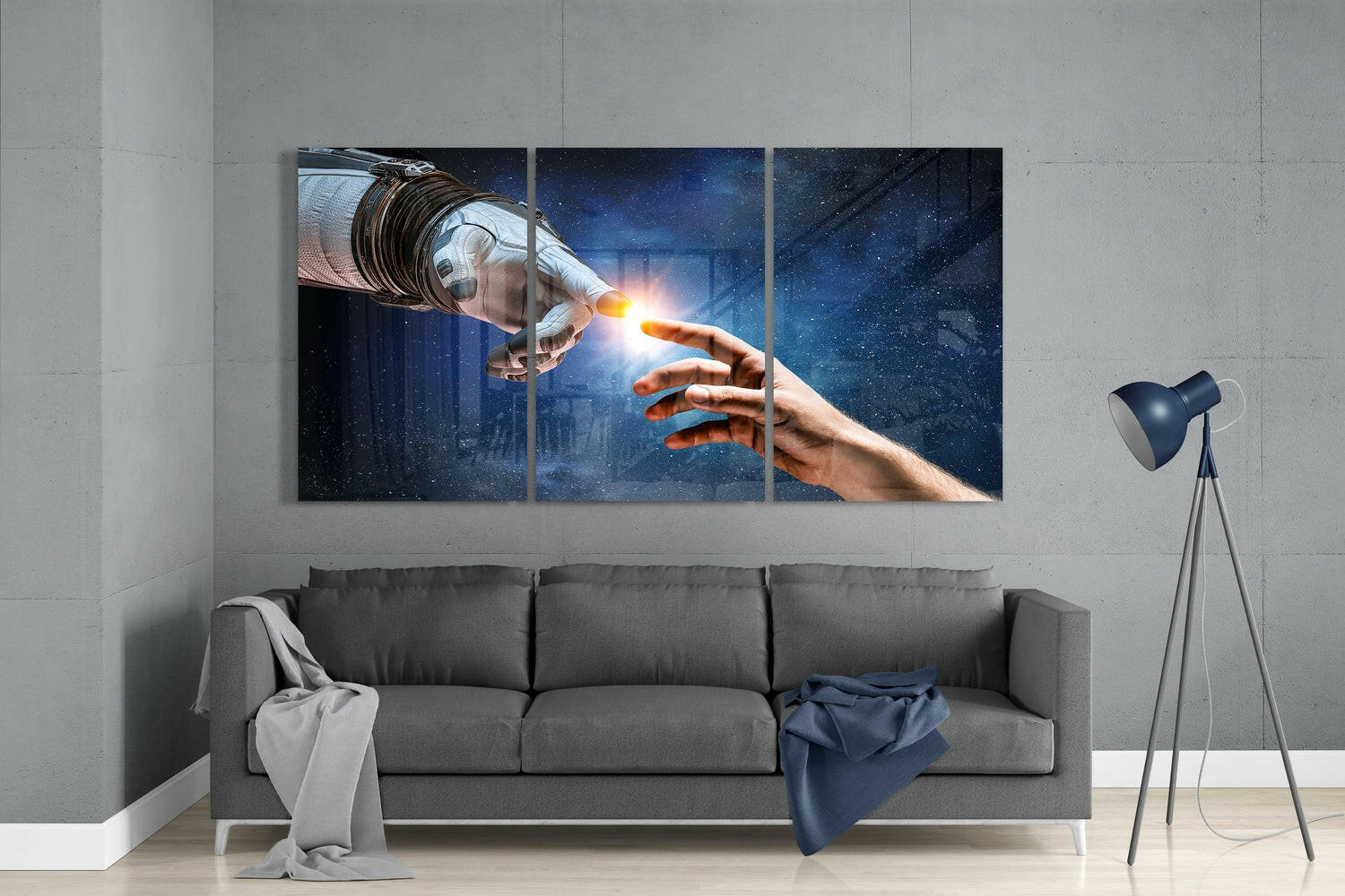 Three-panel glass wall art depicting an astronaut's gloved hand touching a human hand with glowing light, displayed above a gray sofa in a modern interior.