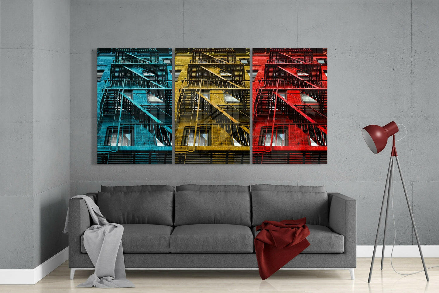 Three glass wall art panels in cyan, yellow, and red display fire escape architecture mounted above a gray sofa in a modern living room.