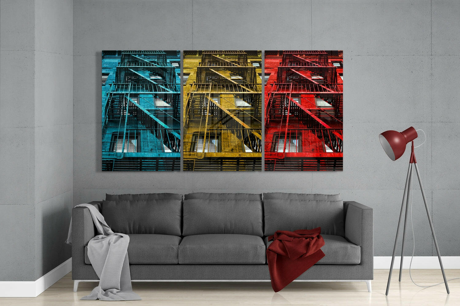 Three glass wall art panels in cyan, yellow, and red display fire escape architecture mounted above a gray sofa in a modern living room.