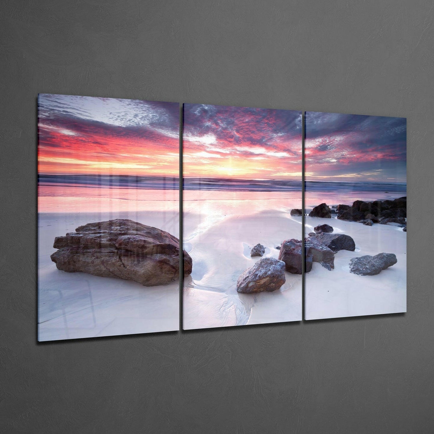 Three-panel glass wall art displaying a beach sunset with rocks in shallow water, vibrant pink and orange sky, mounted on gray wall.