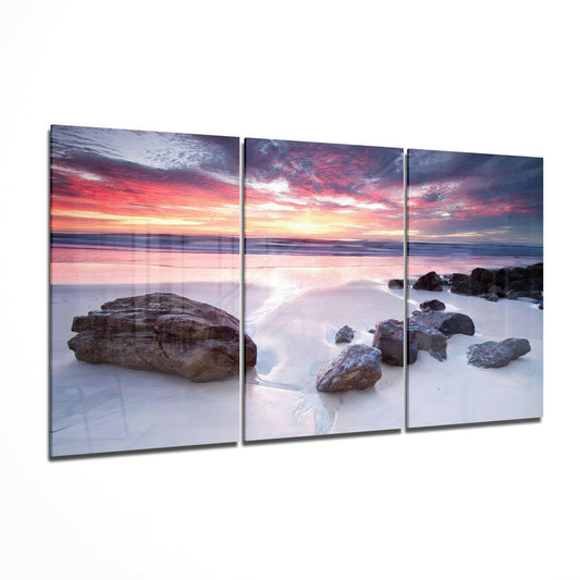 Three-panel glass wall art displaying a rocky beach at sunset with pink and orange sky reflecting on wet sand and shallow water.