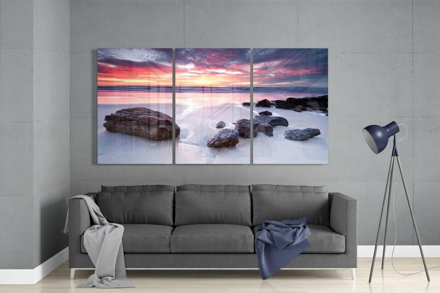 Three-panel glass wall art displaying a rocky beach sunset with pink and orange sky above smooth water and scattered boulders, hung above a gray sofa in a modern living room.
