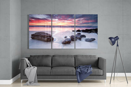 Three-panel glass wall art displaying a rocky beach sunset with pink and orange sky above smooth water and scattered boulders, hung above a gray sofa in a modern living room.