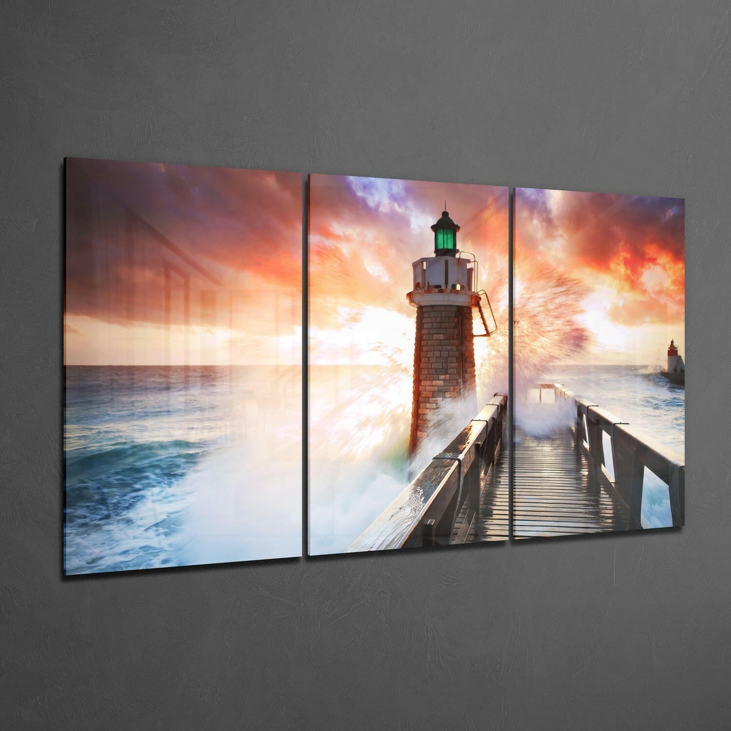 Three-panel glass wall art displaying a lighthouse on a wooden pier with dramatic orange and purple sunset sky and crashing waves.