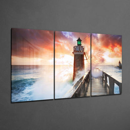 Three-panel glass wall art displaying a lighthouse on a wooden pier with dramatic orange and purple sunset sky and crashing waves.