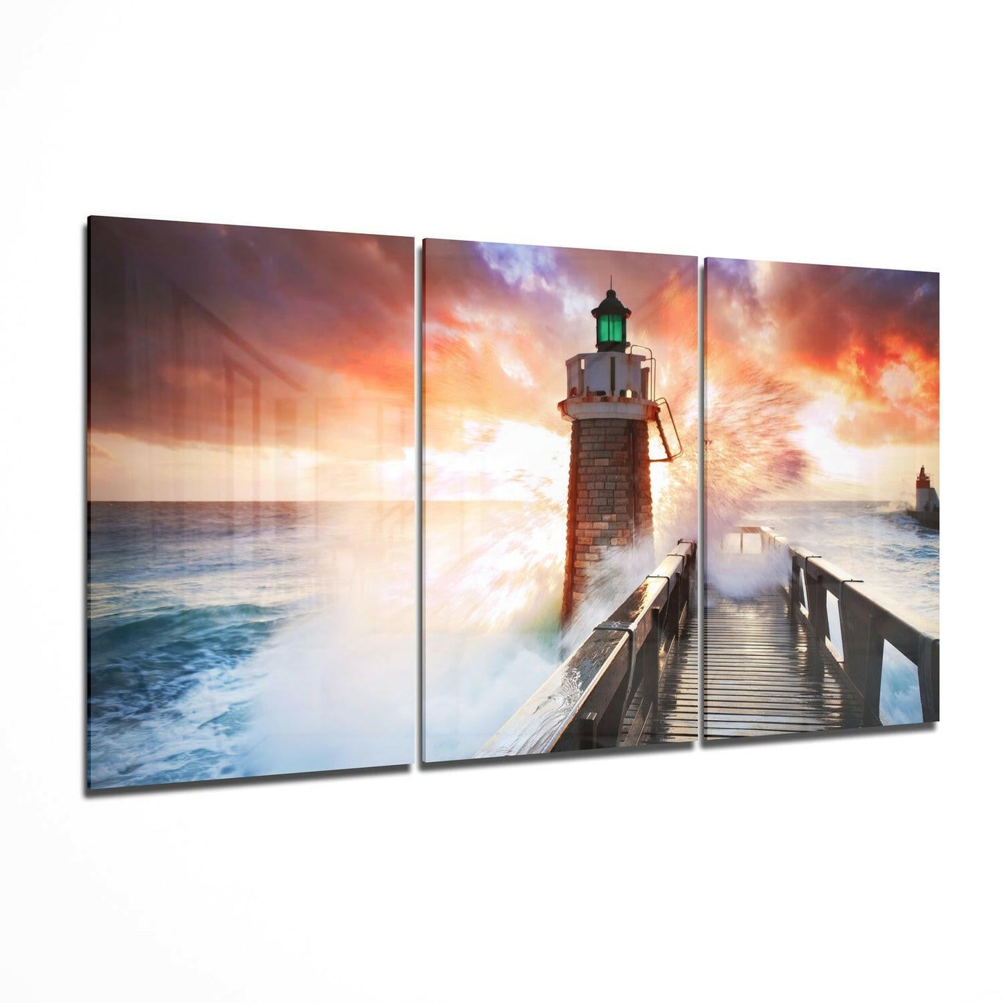 Three-panel glass wall art displaying a lighthouse with green light on a pier during dramatic sunset with crashing waves and orange sky.