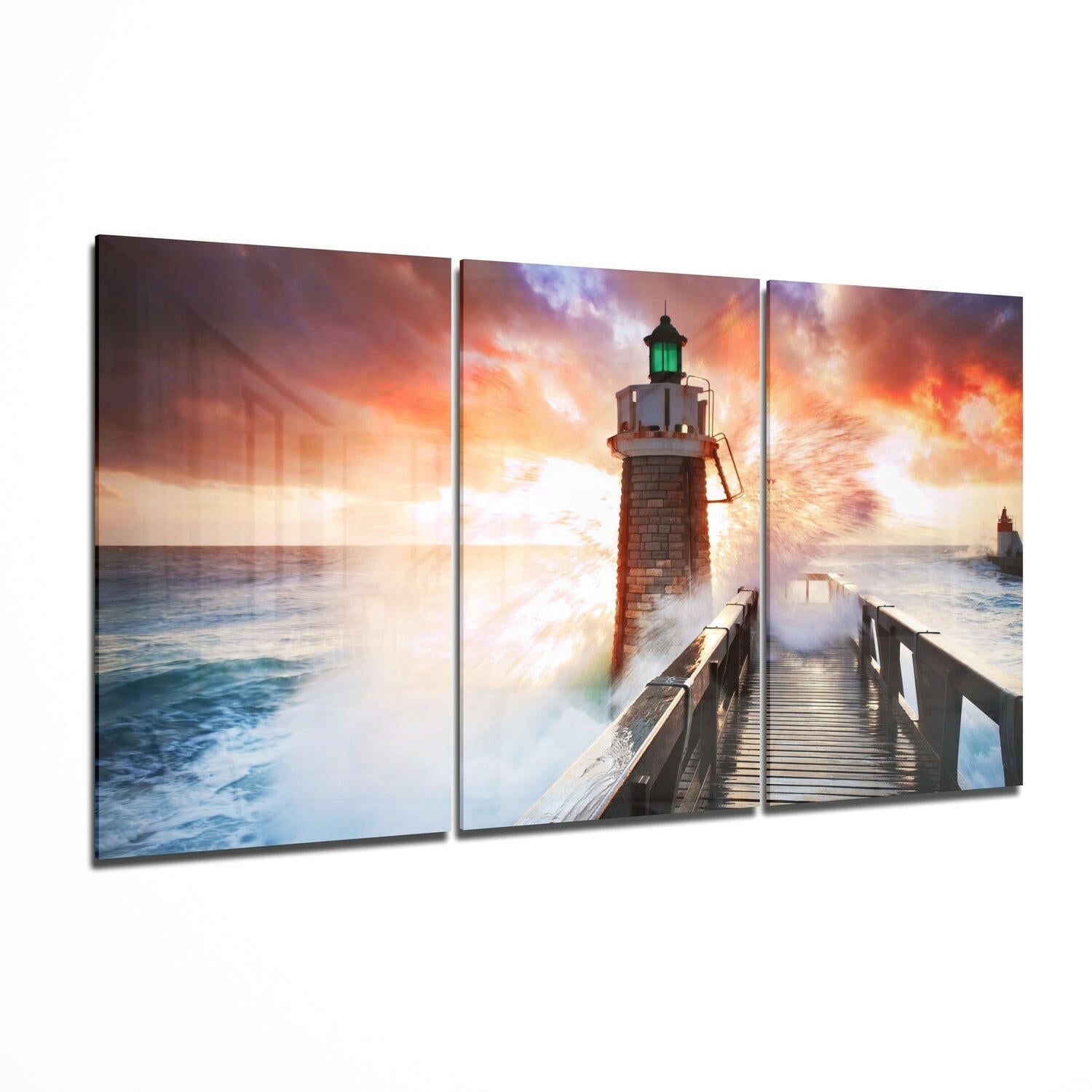 Three-panel glass wall art displaying a lighthouse with green light on a pier during dramatic sunset with crashing waves and orange sky.