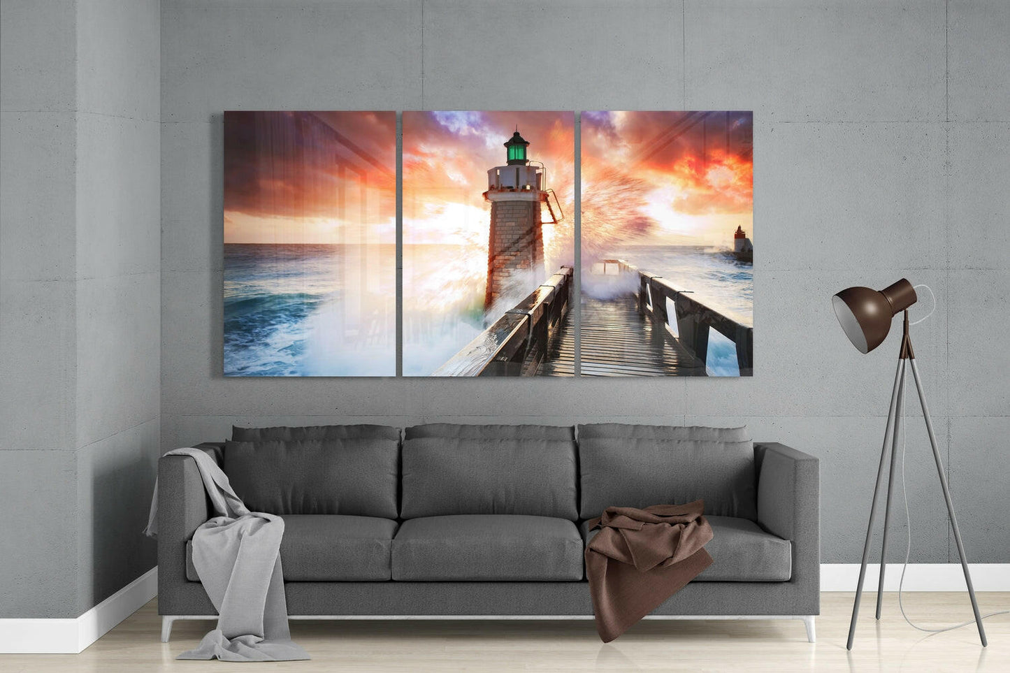 Three-panel glass wall art above gray sofa depicting lighthouse on pier with dramatic sunset sky and ocean waves.