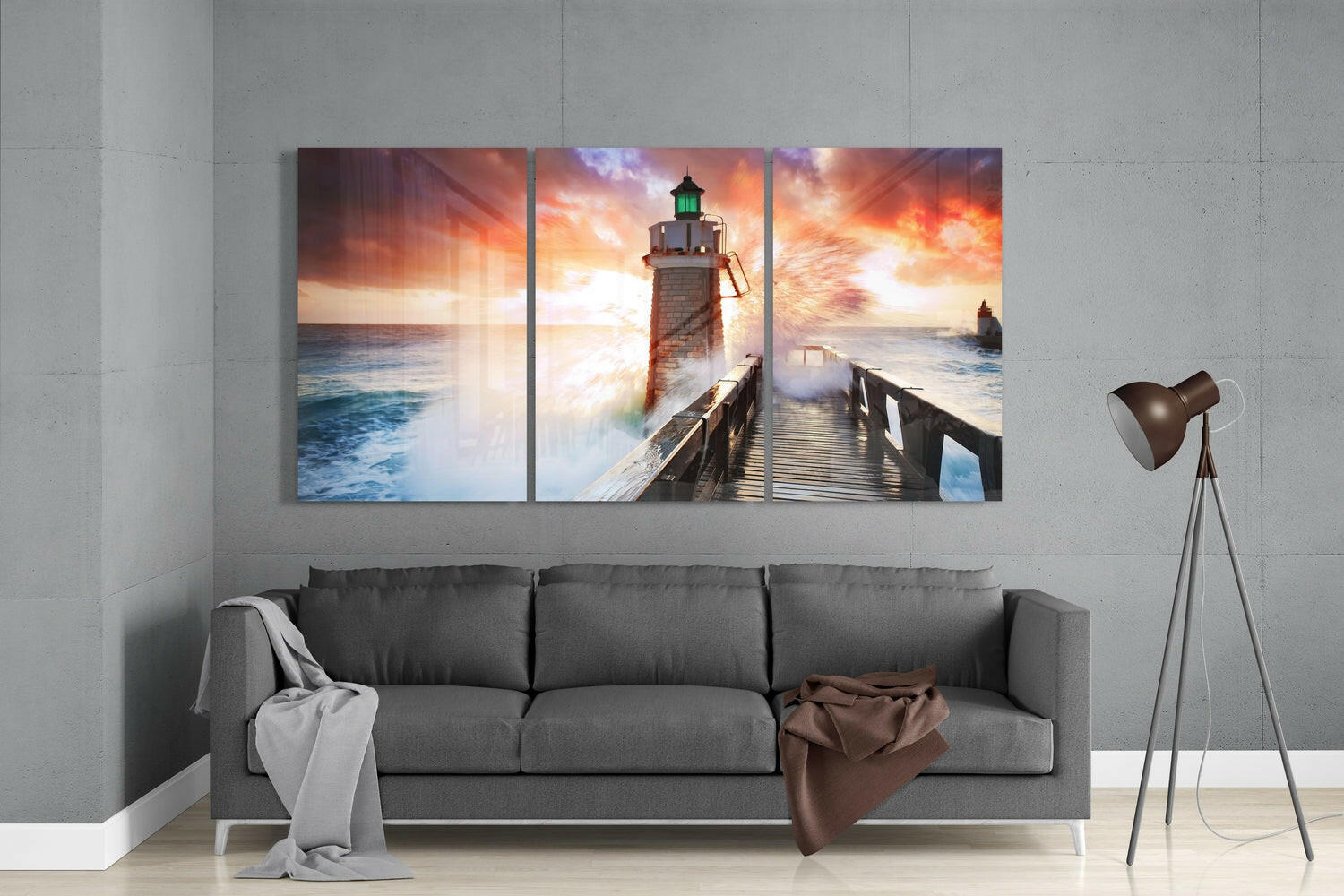 Three-panel glass wall art above gray sofa depicting lighthouse on pier with dramatic sunset sky and ocean waves.