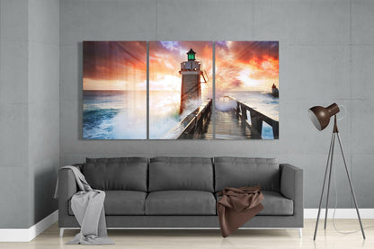 Three-panel glass wall art above gray sofa depicting lighthouse on pier with dramatic sunset sky and ocean waves.
