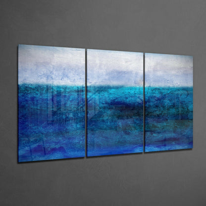 Three-panel blue glass wall art depicting a seascape with layered horizons of sky and ocean gradating from white to deep blue, installed on gray wall.