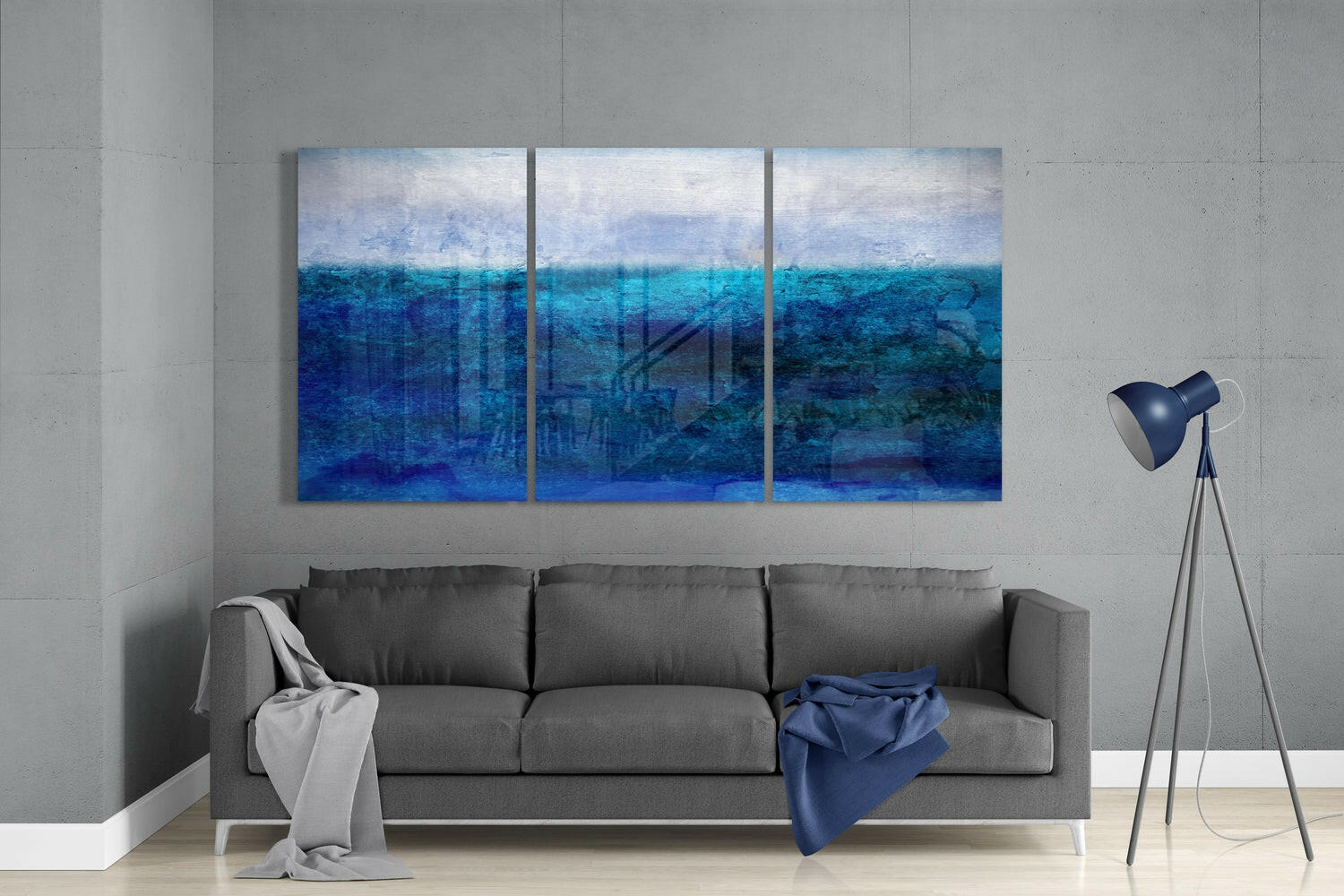 Three-panel blue gradient seascape glass art displayed above gray sofa in minimalist interior setting.