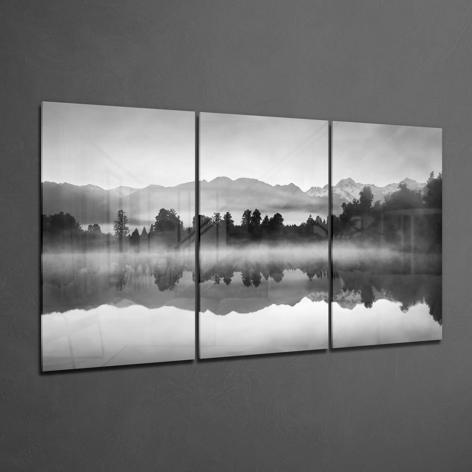 Three-panel black and white glass wall art displaying a misty mountain lake landscape with forested shoreline and mirror reflections.