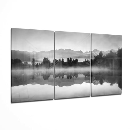 Three-panel black and white glass wall art displaying misty mountains and forested landscape reflected in still water below.