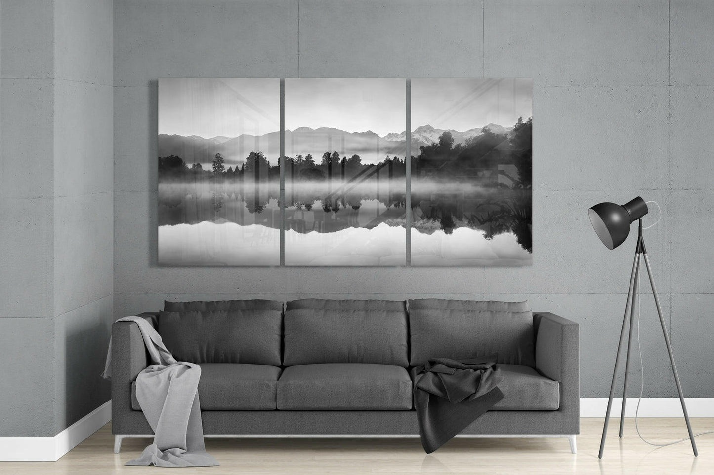 Three-panel black and white glass wall art depicting a misty lake with forested shoreline and mountain reflections, displayed above a gray sofa in a minimalist interior.