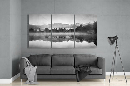 Three-panel black and white glass wall art depicting a misty lake with forested shoreline and mountain reflections, displayed above a gray sofa in a minimalist interior.