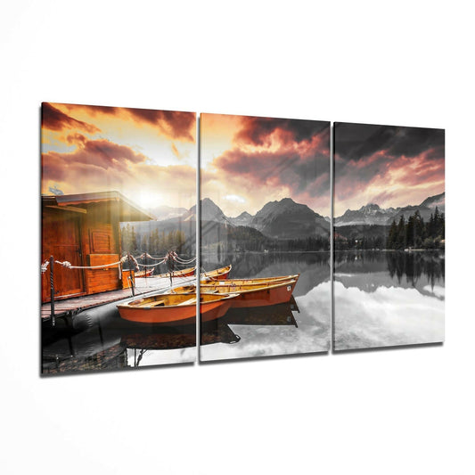 Three-panel glass wall art displaying red boats on a mountain lake at sunset with golden sky and forest reflections.