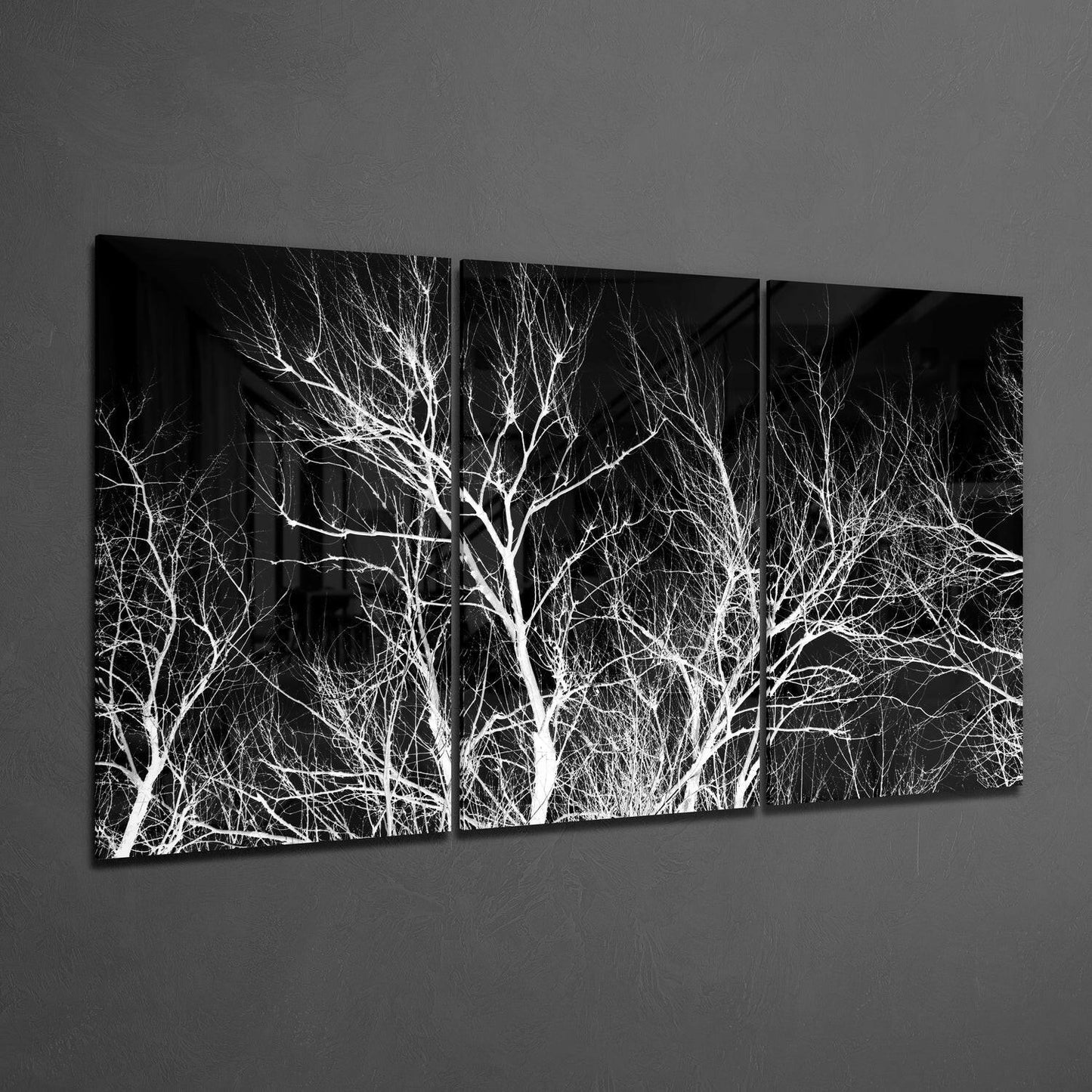 Three-panel black and white glass wall art displaying bare tree branches against dark background, mounted on gray wall.