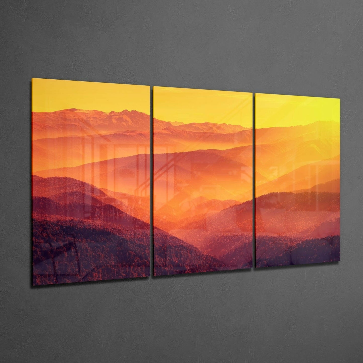 Three-panel glass wall art displaying layered mountains in warm orange and red tones beneath a yellow sky at sunset, mounted on gray wall.