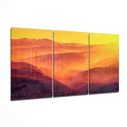 Three-panel glass wall art displaying layered mountains in warm orange and red tones beneath a golden yellow sky at sunset.