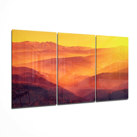 Three-panel glass wall art displaying layered mountains in warm orange and red tones beneath a golden yellow sky at sunset.