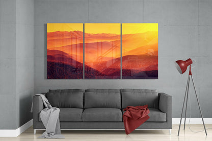 Three-panel glass wall art depicting layered mountains and a golden sunset, displayed above a gray sofa in a modern living room.