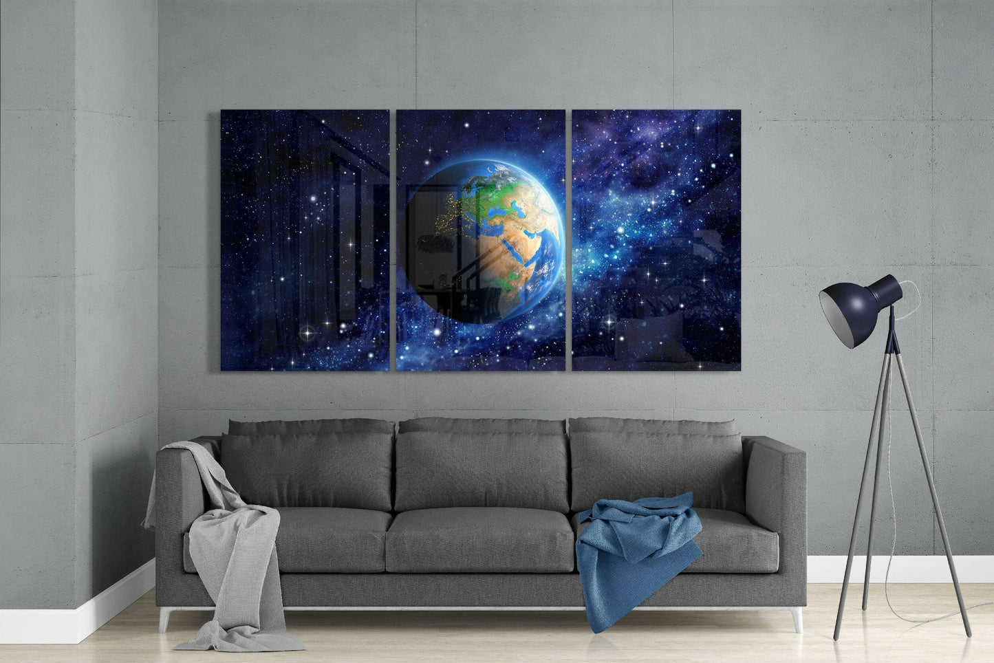 Three-panel glass art displaying Earth against a starry blue galaxy background, hung above a gray sofa in a modern interior.