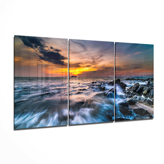 Three-panel glass wall art displaying a rocky coastline at sunset with golden sky, crashing waves, and dark rocks, shown at an angle against white background.