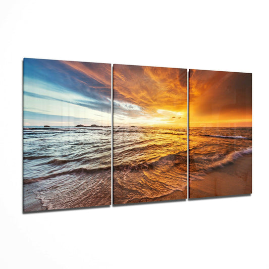 Three-panel glass wall art displaying a beach sunset scene transitioning from cool blue tones on left to golden orange on right, with waves and shoreline visible.