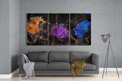 Three abstract glass art panels with orange, purple, and blue brushstrokes on black backgrounds with gold crackle texture, hung above a gray sofa in a modern living room.