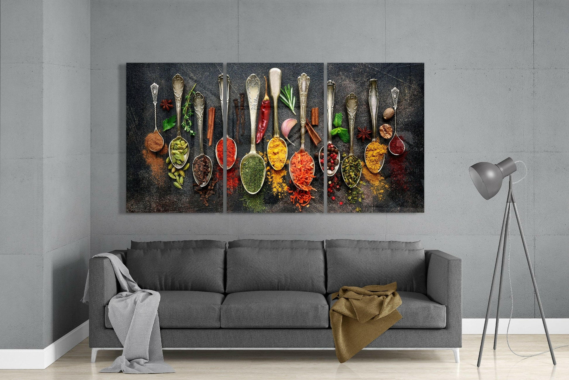 Three-panel glass art displaying ornate spoons filled with colorful spices and fresh herbs on dark textured background, hung above gray sofa in modern living room.