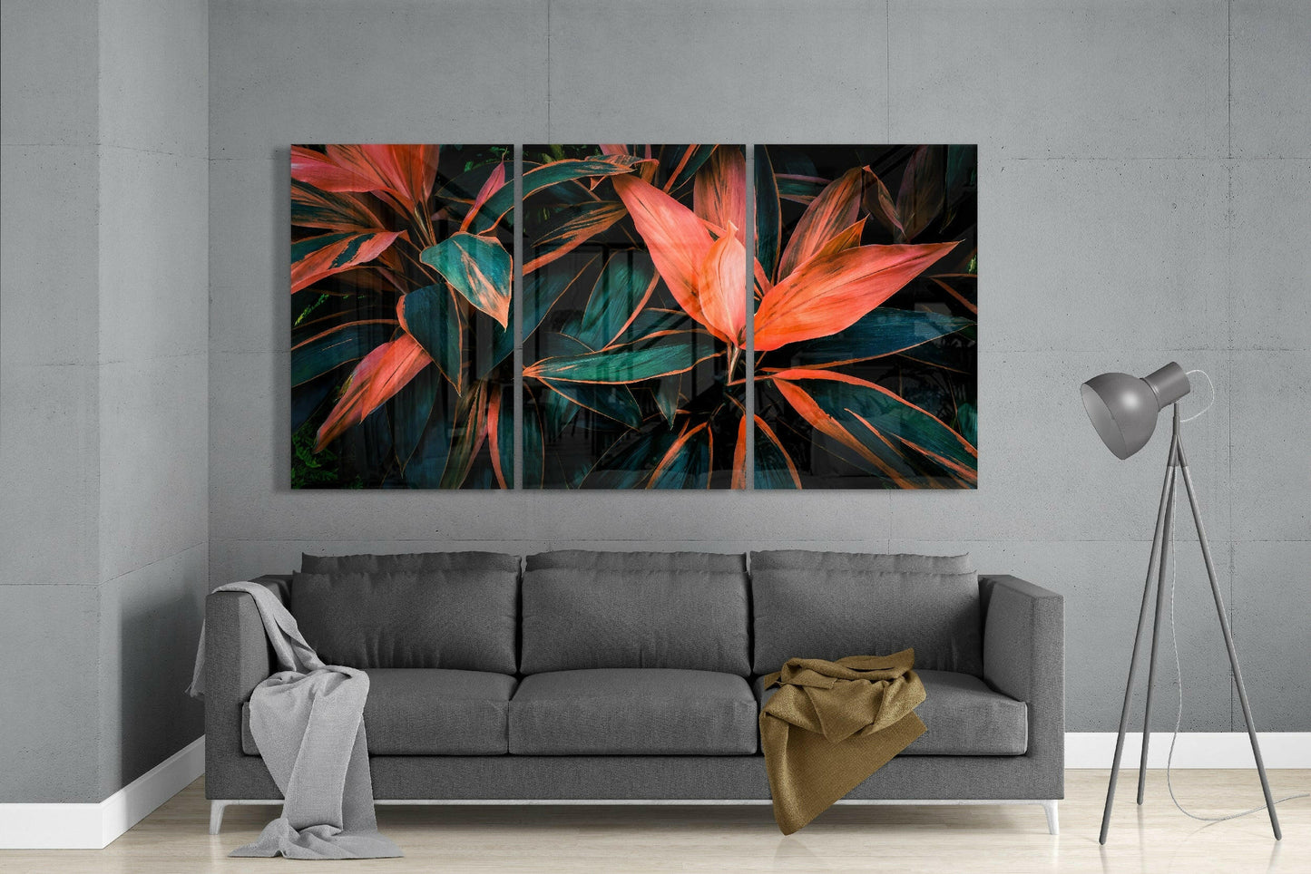 Three-panel glass art displaying vibrant red and orange cordyline leaves with teal foliage against dark background, installed above gray sofa.