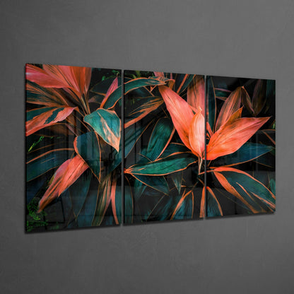 Three-panel glass art display mounted on gray wall showing vibrant red and orange cordyline leaves with teal and dark green foliage against black background.
