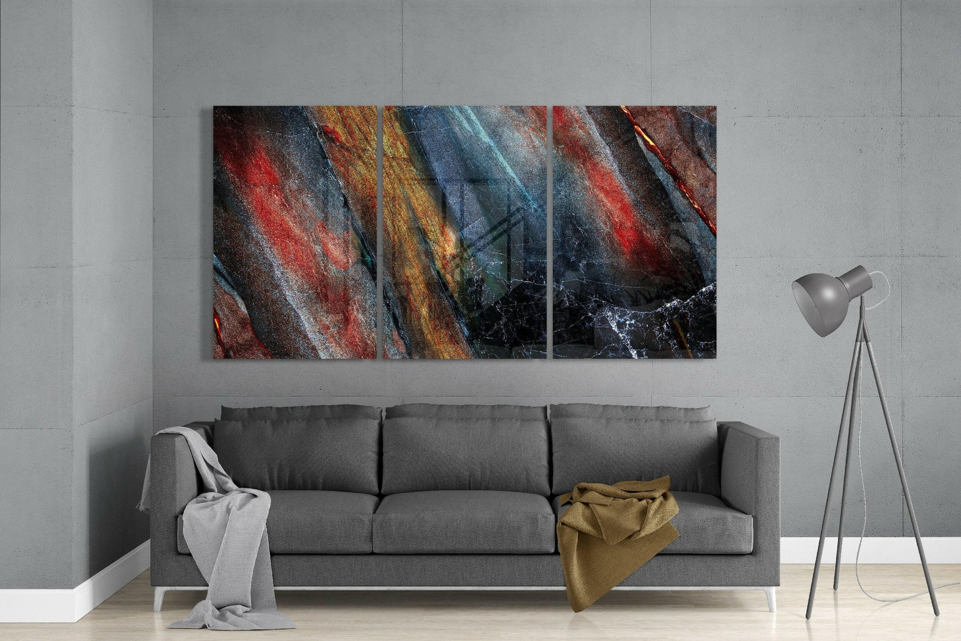 Three-panel abstract glass art with diagonal stripes in red, gold, blue, and black hung above a gray sofa in a modern interior.