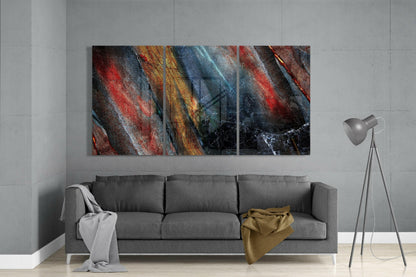 Three-panel abstract glass art with diagonal stripes in red, gold, blue, and black hung above a gray sofa in a modern interior.
