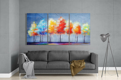 Triptych glass art displaying colorful abstract trees with vibrant red, orange, yellow, and blue foliage above a blue horizon, hung above a gray sofa in a modern interior.