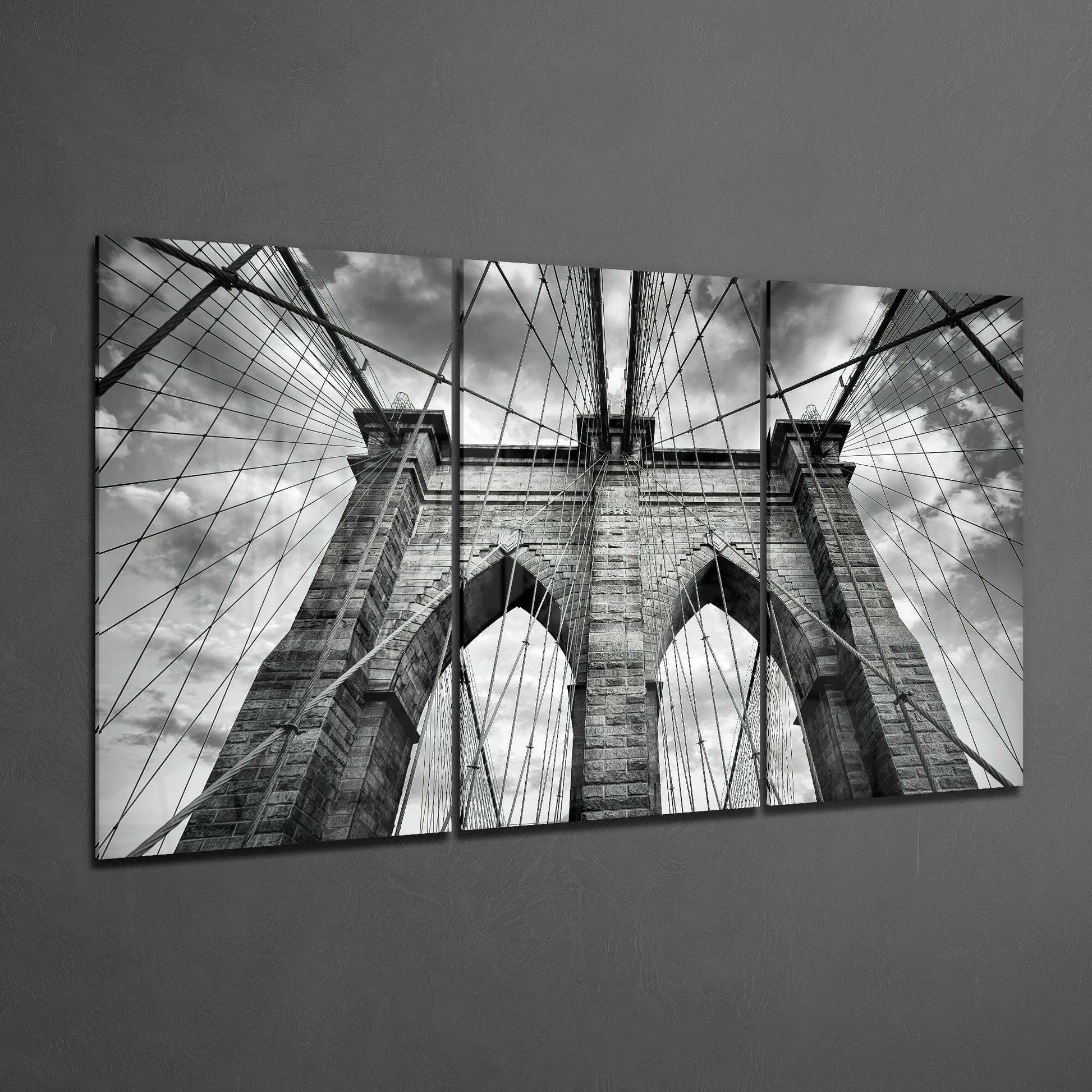 Black and white upward view of Brooklyn Bridge towers and cables against cloudy sky, printed on three glass panels.