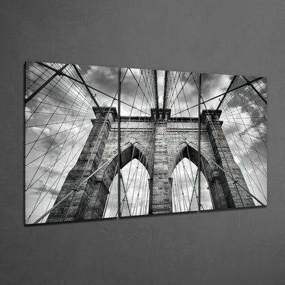 Black and white upward view of Brooklyn Bridge towers and cables against cloudy sky, printed on three glass panels.