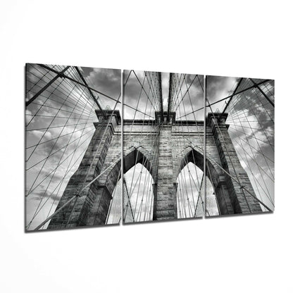 Three-panel black and white glass art displaying upward perspective of Brooklyn Bridge towers, cables, and sky.