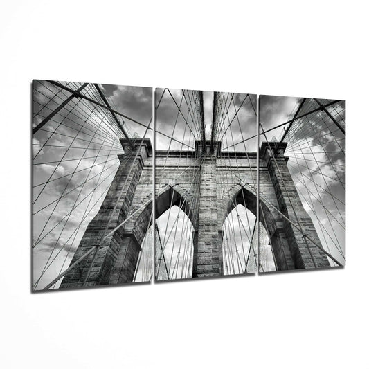 Three-panel black and white glass art displaying upward perspective of Brooklyn Bridge towers, cables, and sky.