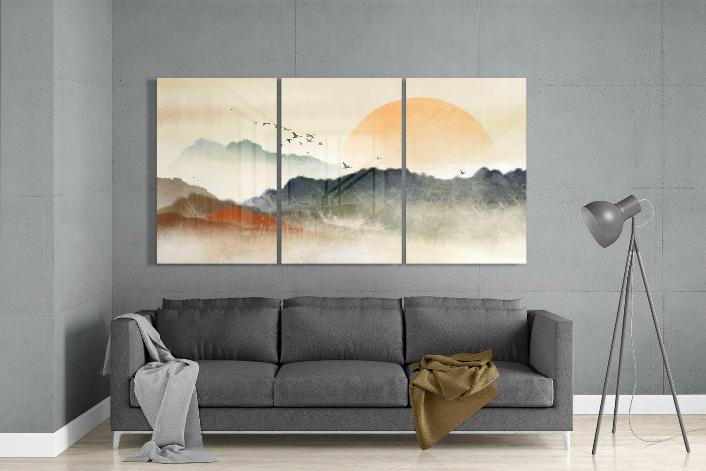 Three-panel glass art depicting misty mountains with large golden sun and flying birds, displayed above gray sofa in modern living room.