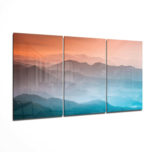 Three-panel glass wall art displaying layered misty mountains transitioning from warm orange tones at top to cool blue tones at bottom.