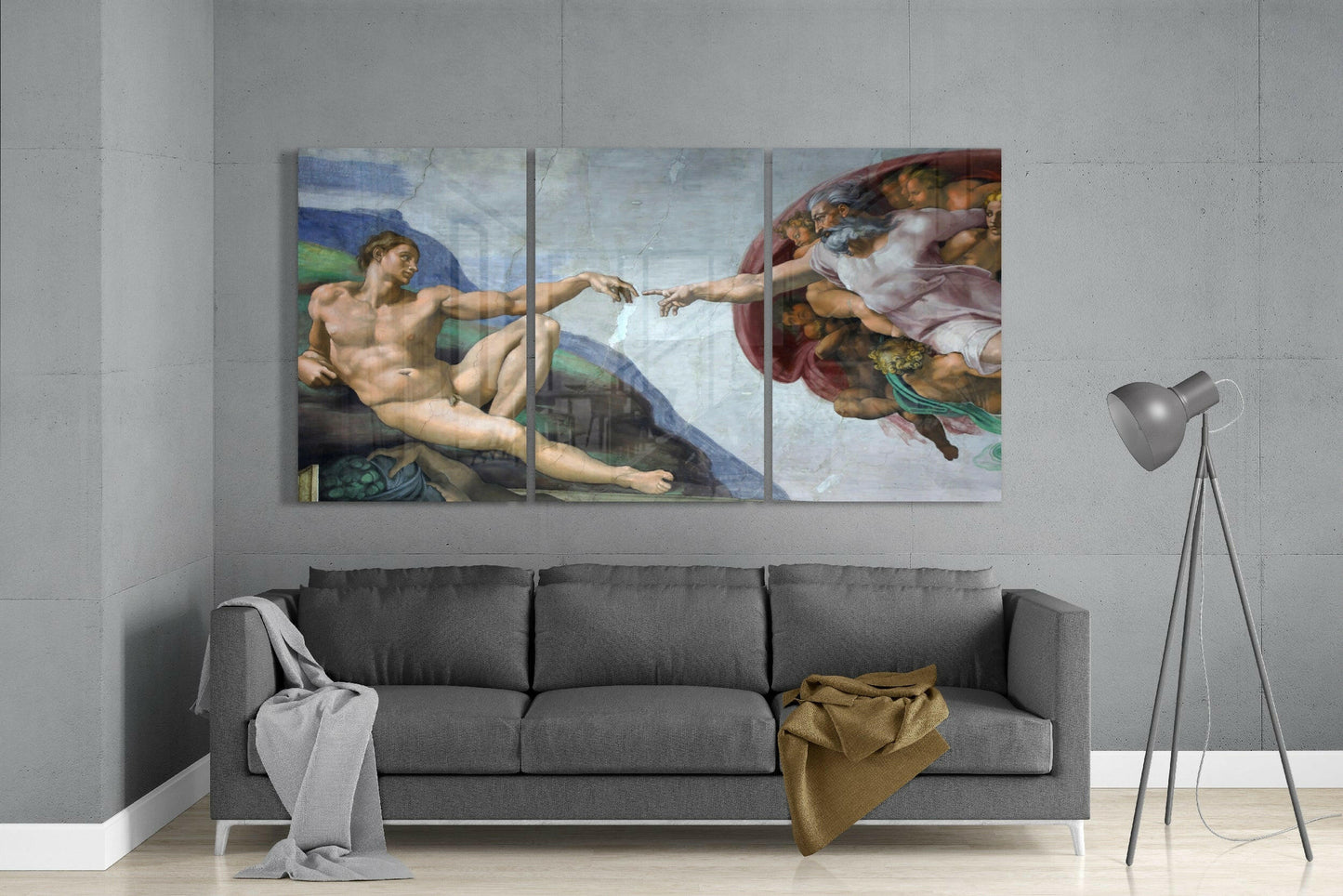 Three-panel glass art print of Michelangelo's Creation of Adam hung above gray sofa in modern living room.