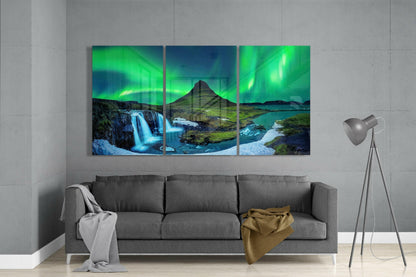 Three-panel glass art displaying Icelandic landscape with green aurora borealis, mountain, waterfall, and turquoise water, hung above gray sofa.