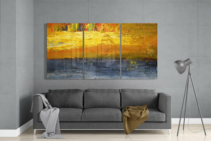 Three-panel abstract yellow glass artwork with orange and blue tones displayed above gray sofa in modern living room.