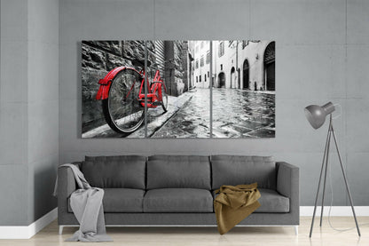 Three-panel glass art mounted above gray sofa displaying red bicycle on cobblestone street with monochrome buildings.
